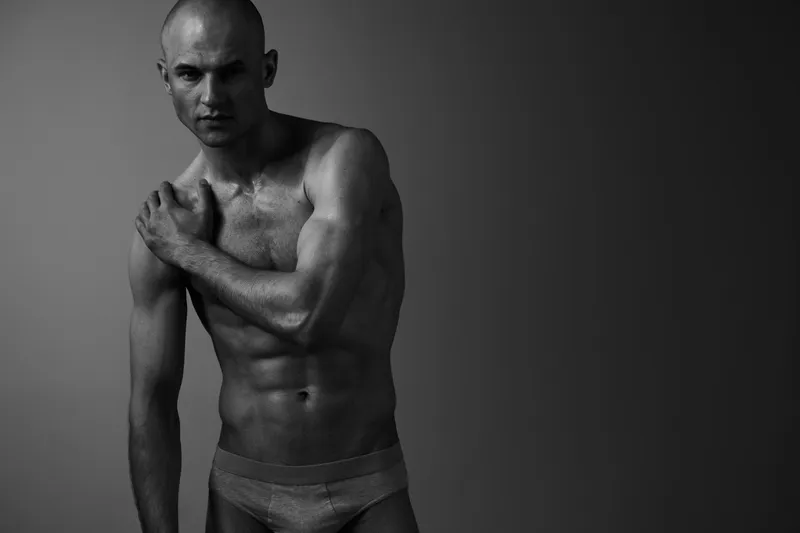 Black and white image of a muscular man posing gracefully, showcasing strong physique and artistry in a dramatic light. Conveying strength and emotion effectively.