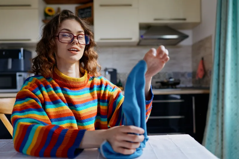 Woman in a colorful striped sweater sits at a home table in a bright kitchen, smiling while folding laundry, a warm casual lifestyle moment captured indoors