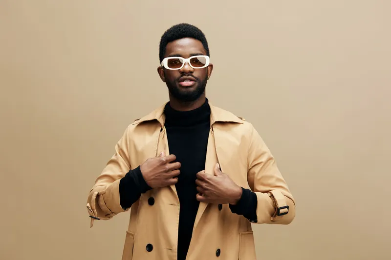 Stylish young man in a beige trench coat and white sunglasses poses confidently against a neutral background, showcasing modern fashion and individuality