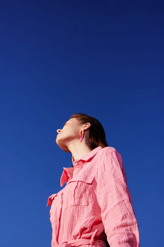 A young woman with light skin gazes upward against a clear blue sky, wearing a pink shirt that reflects a casual summer vibe and conveys a sense of freedom and aspiration