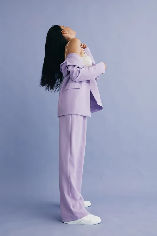 woman in lilac suit stands with head tilted back, posing in front of purple studio backdrop, wearing white sneakers and light top, showcasing modern fashion style