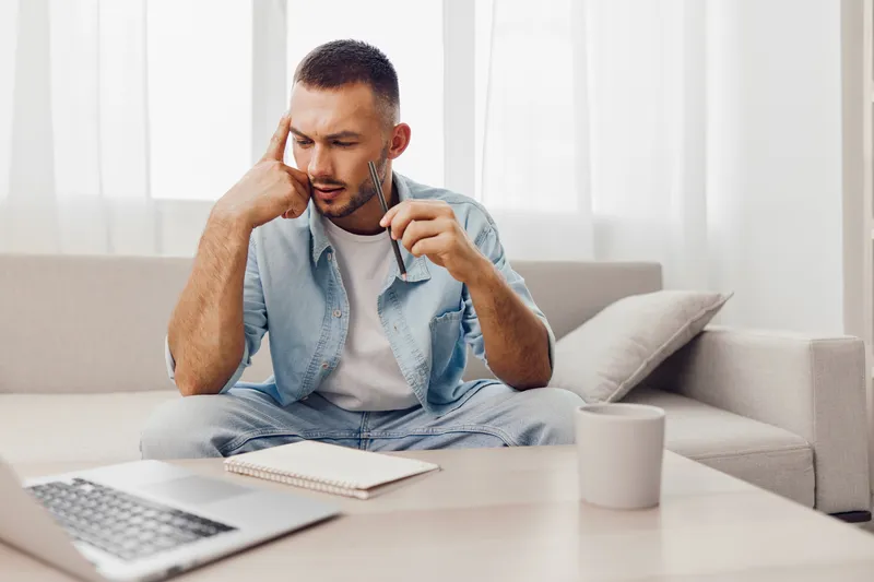 Thoughtful man sitting at home with a laptop, analyzing data and taking notes while enjoying coffee. Ideal for work-from-home themes and productivity concepts.