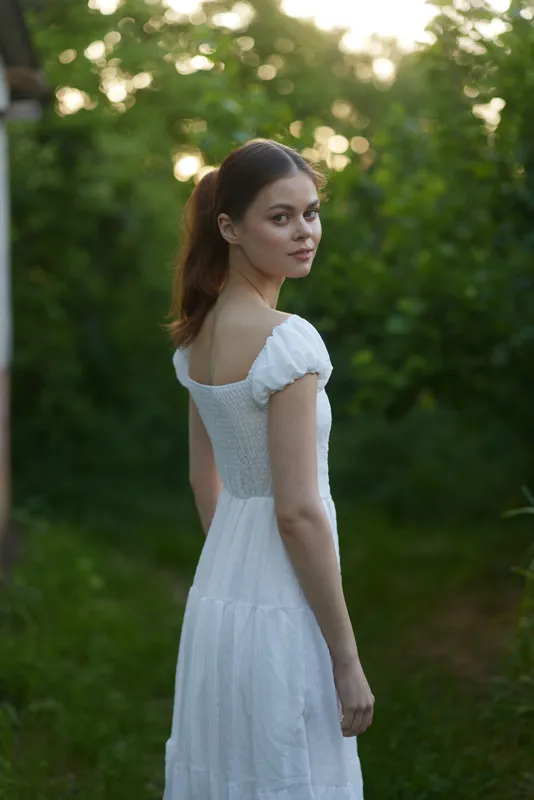 Young woman in a white dress standing in a green garden, exuding calmness and beauty, surrounded by nature Ideal for topics on summer fashion and serenity