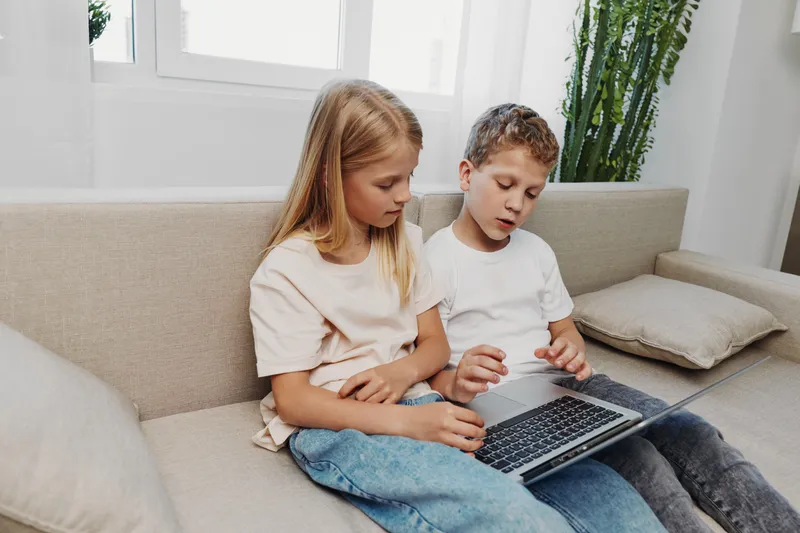 Two children sitting on sofa using laptop together, focused on screen in bright living room. Concept of learning, technology, and childhood interaction.