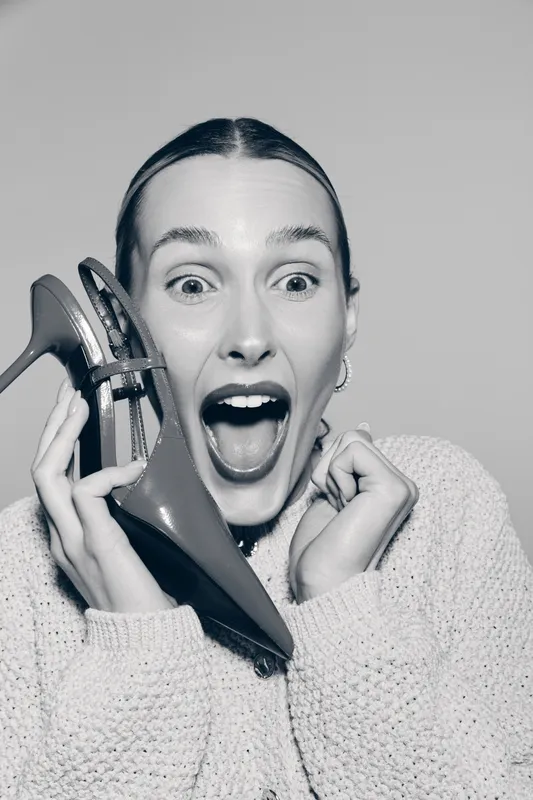 Caucasian woman with light skin and red hair holds high heel shoe near face, expressing surprise and excitement. Studio portrait in monochrome with casual sweater. Concept for fashion and emotions.