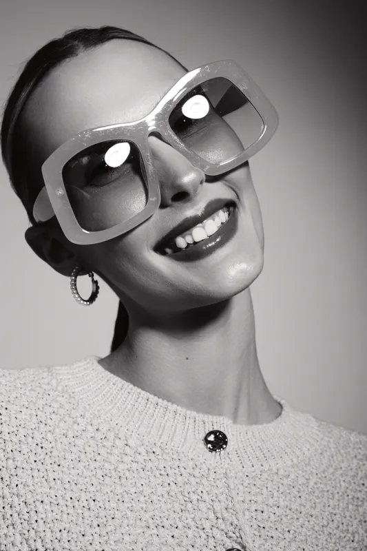 Stylish Caucasian woman with pale skin wearing oversized sunglasses and hoop earrings smiles confidently against a plain background in black and white portrait.