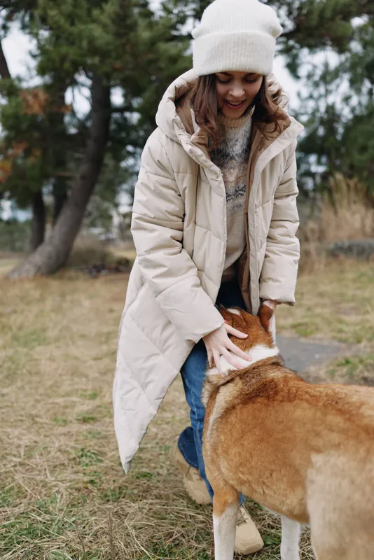 Woman, dog, winter, outdoor, coat, park, interaction, warmth, happy, fashion. A joyful scene shows a woman in a padded coat and beanie greeting a dog in a grassy park with trees and calm weather