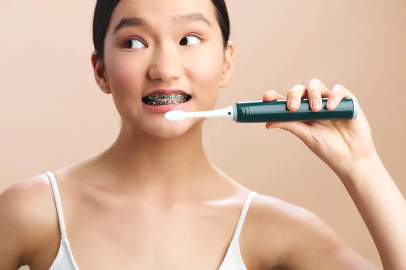 Asian woman brushes her teeth with an electric toothbrush in a bright, minimal setting, conveying personal care and wellness. Her calm focus highlights daily hygiene in a clean, modern routine.