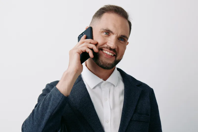 A confident male professional on a call, dressed in a smart blazer and white shirt, stands against a clean solid color background, conveying focus, leadership, and effective business communication.