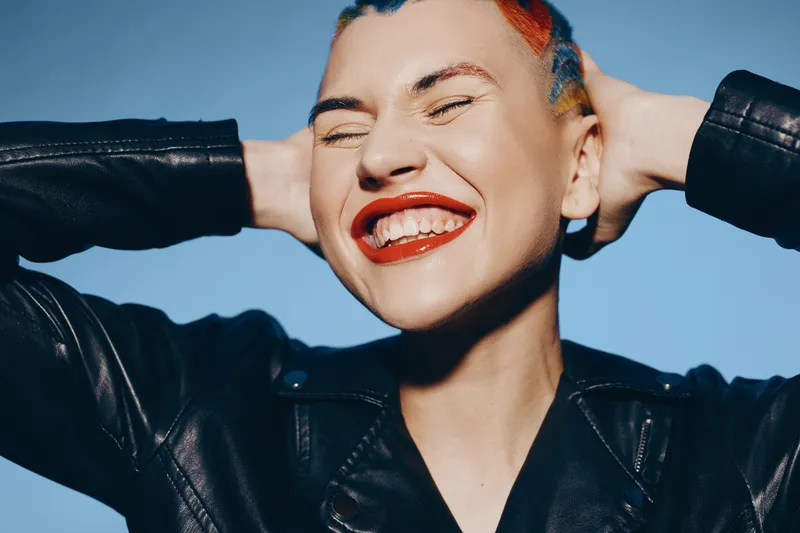 Young woman with colorful short hair smiling broadly, wearing black leather jacket against clear blue sky, expressing joy and carefree attitude.