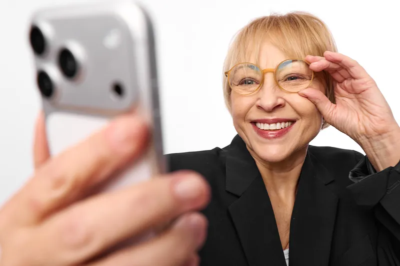 Elderly woman selfie with a smartphone, smile on her face, adjusting glasses with one hand, close portrait on bright background, friendly mood, technology communication