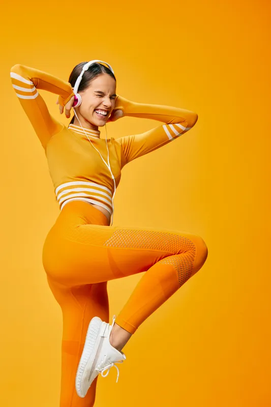 Woman fitness sport in a bright orange outfit, mid jump with headphones and white sneakers, posed against a warm orange backdrop, conveying dynamic movement and energetic workout.