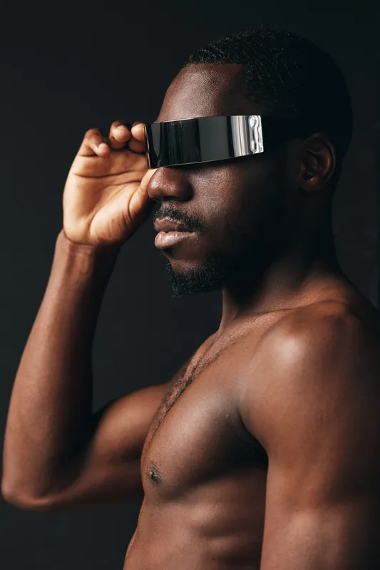 Creative conceptual portrait of a shirtless black man wearing futuristic reflective visor glasses in a visual style with dark background and strong lighting contrast.