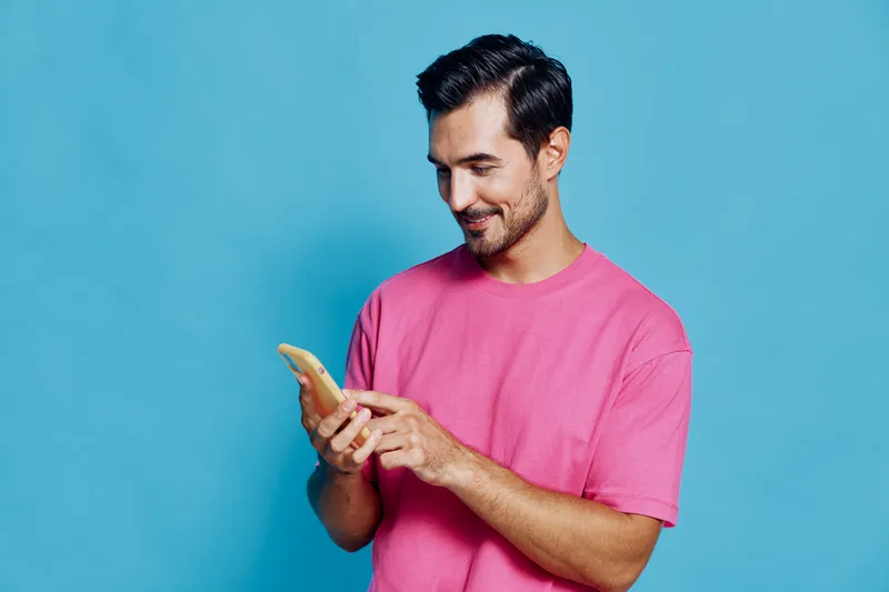 Smiling man in a pink t-shirt looking at his smartphone with a cheerful expression against a bright blue background, engaged in digital communication.