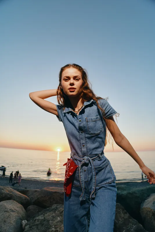 young woman in denim outfit poses near rocky beach at sunset with calm sea and warm sky in background, casual summer fashion and relaxed mood