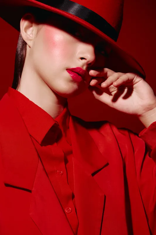 Close-up of elegant woman wearing bright red hat and jacket, red lipstick and glossy glowing skin. Bold makeup and fashion creative in red studio. Concept of beauty and style.
