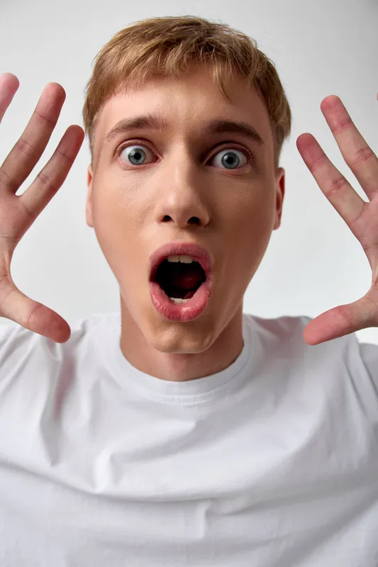 Surprised face with wide eyes and open mouth, hands raised, white tshirt, studio lighting, expressive closeup of a young male in shock, isolated on neutral background