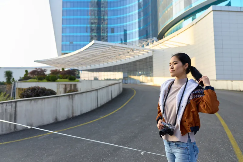 Photography of a woman with camera on an urban rooftop, casual jacket, sunlit cityscape, modern buildings and skyline, street scene conveying travel vitality and creative storytelling