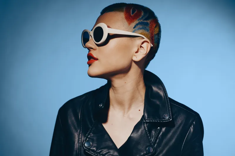 Bold woman with colorful tattoo hairstyle and round white sunglasses wearing black leather jacket, posing confidently against smooth blue background in studio setting.
