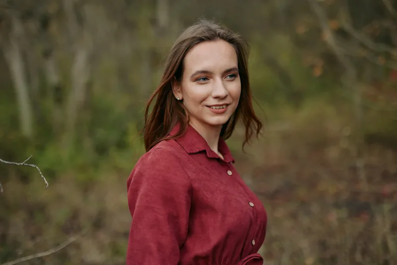 Young woman in a natural woodland portrait, smiling gently with relaxed posture and direct gaze, conveying authenticity and trustworthiness in a candid, documentary style image.