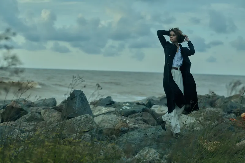 Powerful wind sweeps across a rocky coastline as a woman in a long coat moves dynamically, conveying drama and freedom amid sea spray and overcast clouds