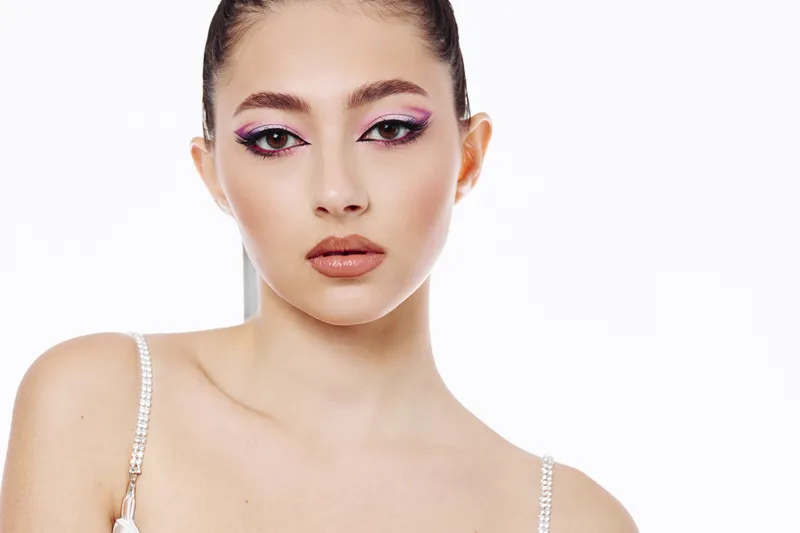 Young woman with creative fashion makeup and glitter eyeliner poses against white background. Concept of beauty, cosmetics, and bold style with artistic flair in a studio setting.