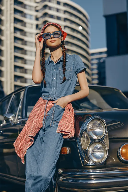 Lifestyle woman in modern boho-western denim street style poses confidently with sunglasses and braided hair by vintage car, captured with warm film color tones.