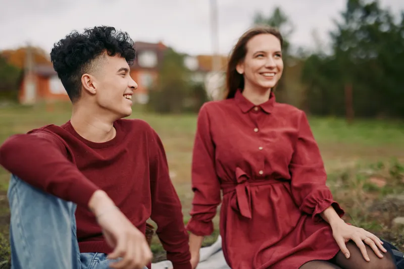 A young couple sits on a picnic blanket, sharing candid smiles and relaxed conversation in a natural outdoor scene, conveying authentic warmth, connection and honest emotion, quietly real.