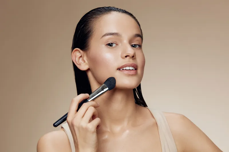 Young woman applying makeup with a foundation brush, showcasing a natural beauty look against a warm, neutral background