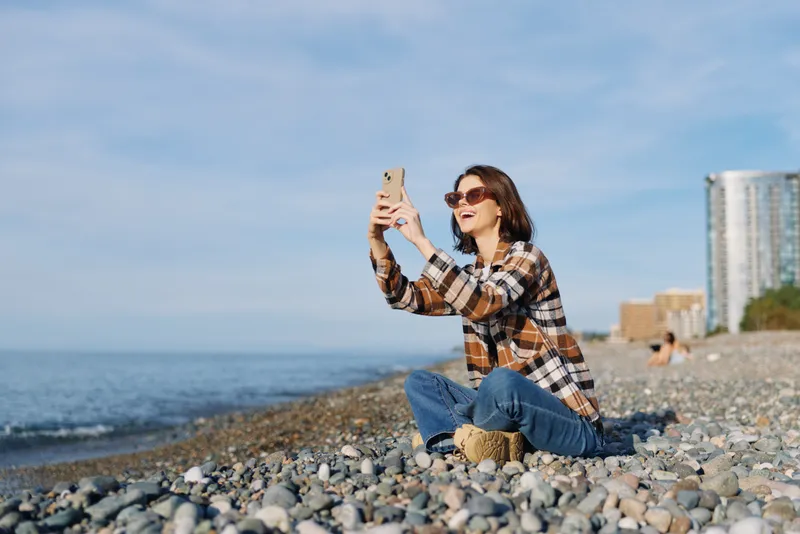 Woman on a pebbled beach, wearing a plaid shirt and jeans, takes a selfie with her phone as the ocean and blue sky frame a relaxed outdoor moment.