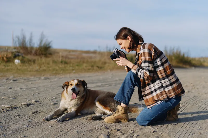Photographer kneeling beside a dog in an open outdoor setting, capturing moments with a handheld camera as the duo explores a dusty landscape under clear skies