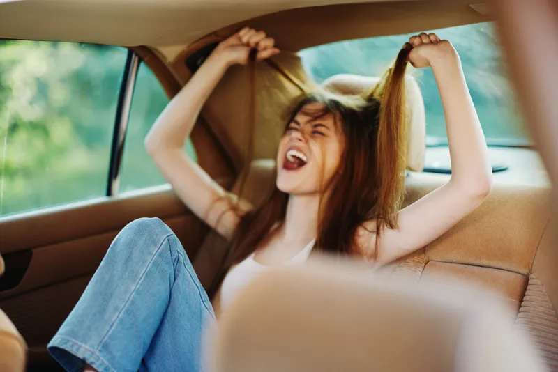 Young woman expressing frustration while pulling her hair in a car, surrounded by a lush green background highlighting her emotions and casual style.