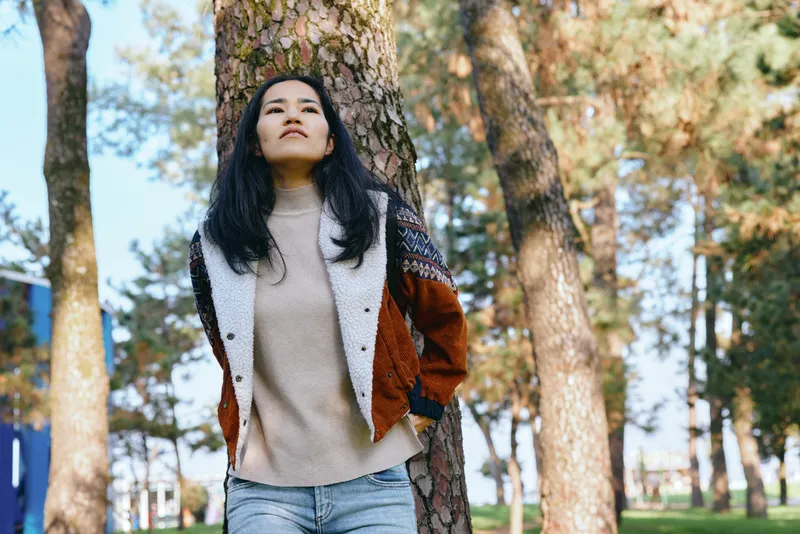 Outdoor scene with a woman leaning against a tree in a park, wearing a chunky sweater and colorblocked jacket, casual autumn fashion captured in bright natural light and calm surroundings