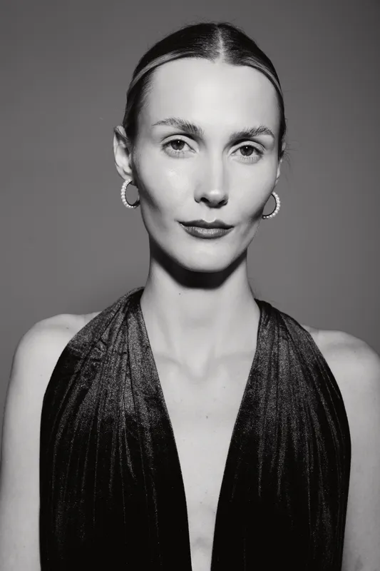 Portrait of a white young woman with light skin and slicked back hair wearing hoop earrings and a dark sleeveless top. Studio black and white photo with confident mood.