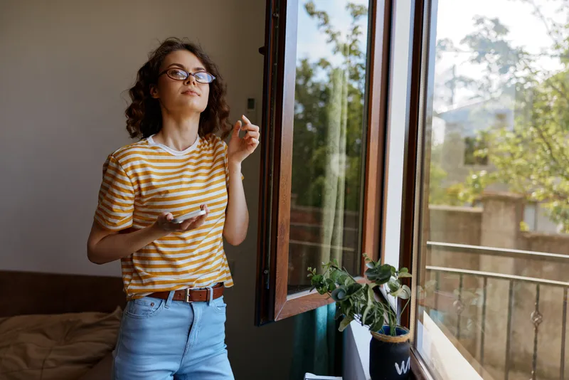 Cheerful woman at home, standing by an open window, sunlight on striped shirt, relaxed posture, observing outdoors, cozy interior, fresh air, natural colors, plant nearby