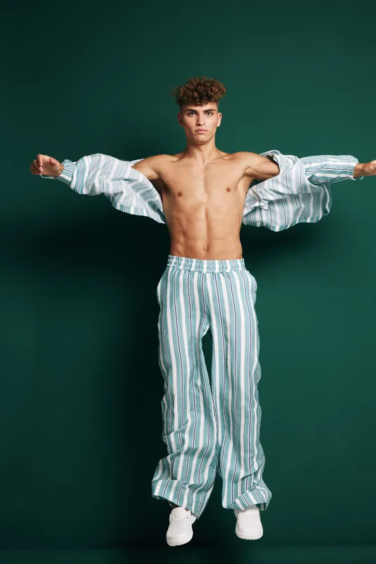 Young shirtless man with athletic body wearing striped pajamas and white sneakers standing with arms extended against dark background isolated in studio portrait.