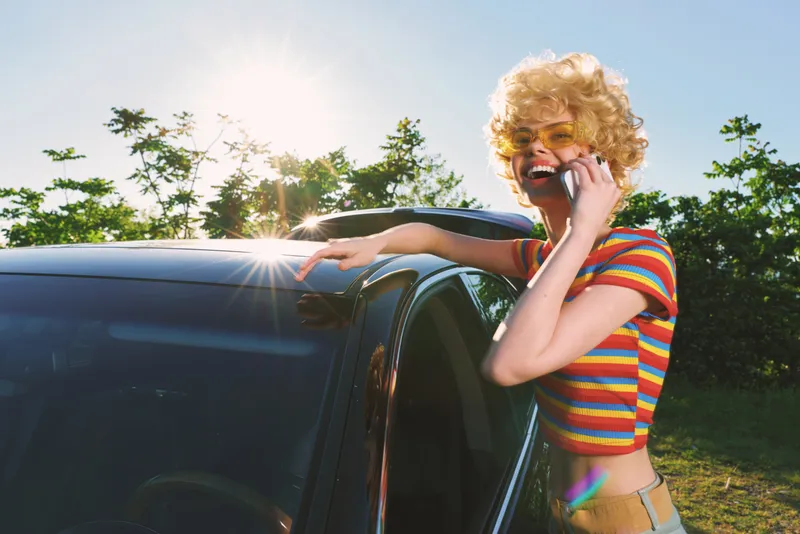 Woman with retro striped shirt talks on a phone beside a car under bright sun, capturing a casual lifestyle moment with vintage film color tones and sunny mood.