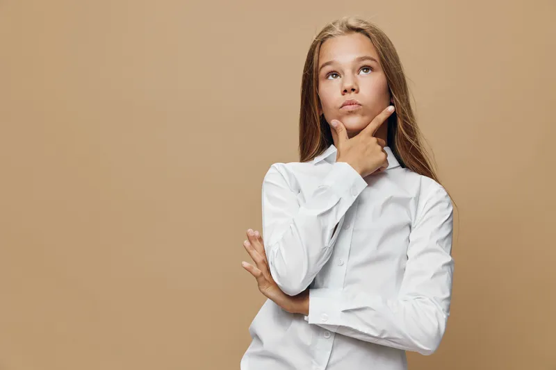 Girl in white shirt standing with hand on chin and thoughtful expression, isolated on beige background for contemplation and decision making concept.