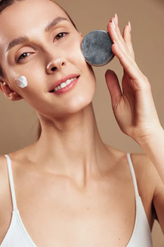 Young woman with clear skin applying cream on face holding cosmetic container close to cheek smiling gently natural beauty skincare routine neutral background