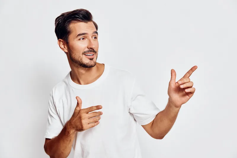 Smiling man wearing casual white t-shirt gestures confidently while looking at the camera, showcasing cheerful expression against a plain background.