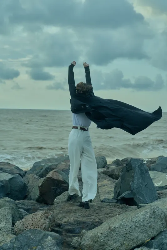 Man standing on rocky shoreline with cape billowing, facing the ocean, embodying freedom, resilience and bold determination against rough seas