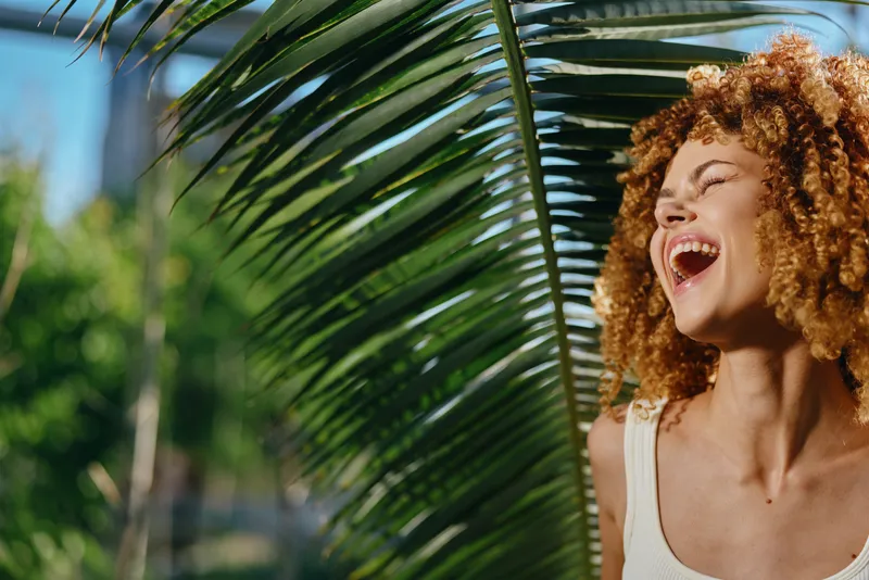 laughing woman with curly hair enjoys sunny day near green tropical palm leaves outdoors in warm natural light, expressing happiness and freedom