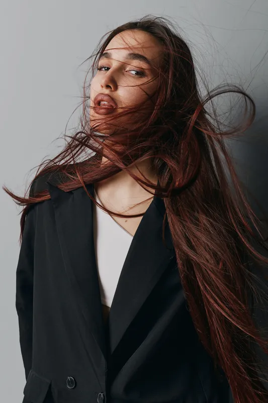 Woman with long hair wearing black blazer posing against plain background, hair flowing dynamically, natural makeup, casual fashion style, portrait with confident expression.