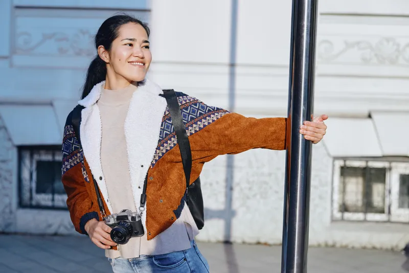 Photography, outdoor, camera, smiling, jacket, backpack, urban, street scene, woman poses with camera in hand beside a pole, enjoying travel and everyday exploration
