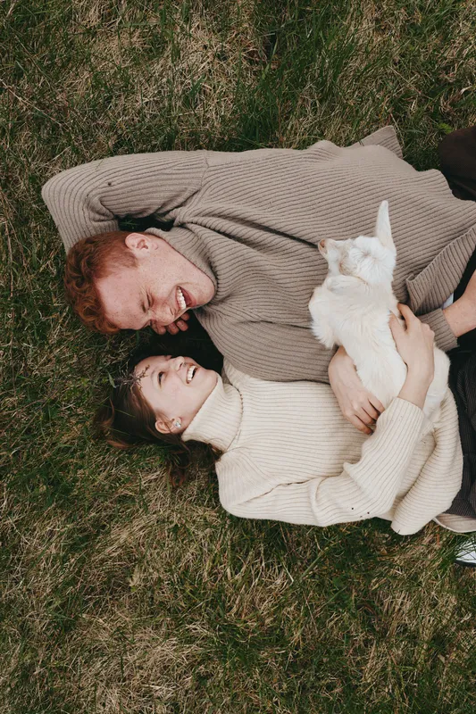 Candid couple outdoors, lying on grass with a goat kid nearby, warm sweaters, relaxed smiles, gentle breeze, sunlit mood, natural tones, lifestyle moment, connection and comfort