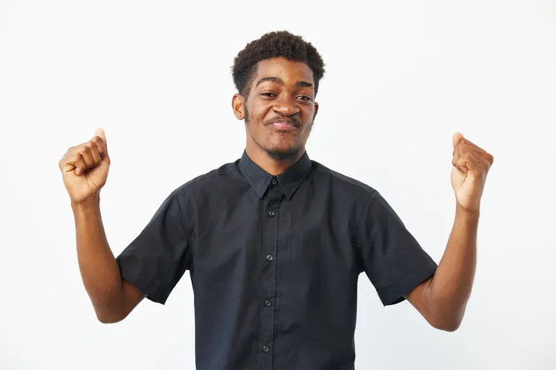 Happy African American man celebrating success with raised fists, wearing a black shirt against a white background. Joyful expression and positive vibes evident.