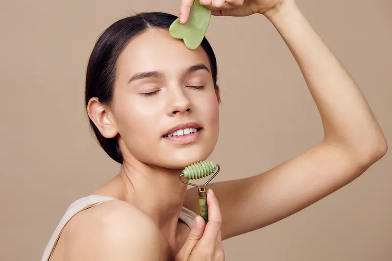 Young woman using a jade roller for facial massage, promoting relaxation and skincare, featuring a minimalist beige background and a soothing expression