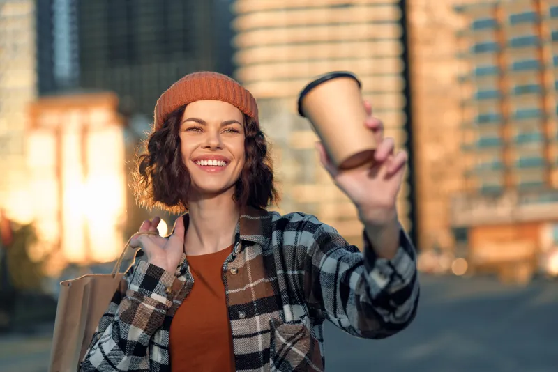 woman coffee smile urban candid golden: young woman in beanie offers a takeaway cup to camera with warm grin during city golden hour, authentic mindful living and emotional storytelling moment