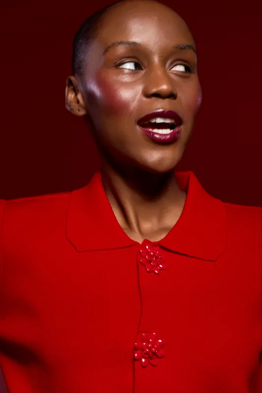 Portrait of a smiling African woman with dark skin wearing a red jacket on a red background. The image highlights fashion, beauty, and confident expression in studio lighting.