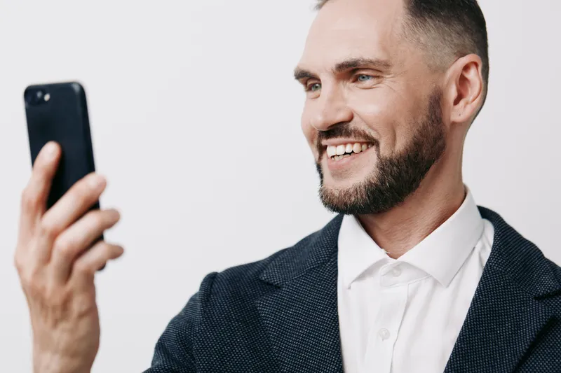 A professional man in a suit smiles while looking at a smartphone, conveying confidence, engagement, and optimism. Isolated light gray colored background enhances focus.